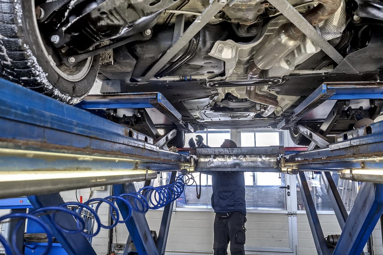 Blick von unten auf ein Auto auf der Hebebühne, darunter ein Prüfer bei der Untersuchung