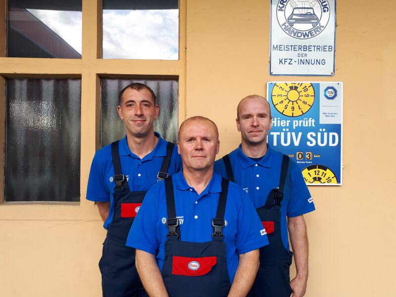 Die drei Mechaniker der Werkstatt in Esso-Latzhosen vor dem Werkstattgebäude, daneben das Schild „Hier prüft TÜV Süd“